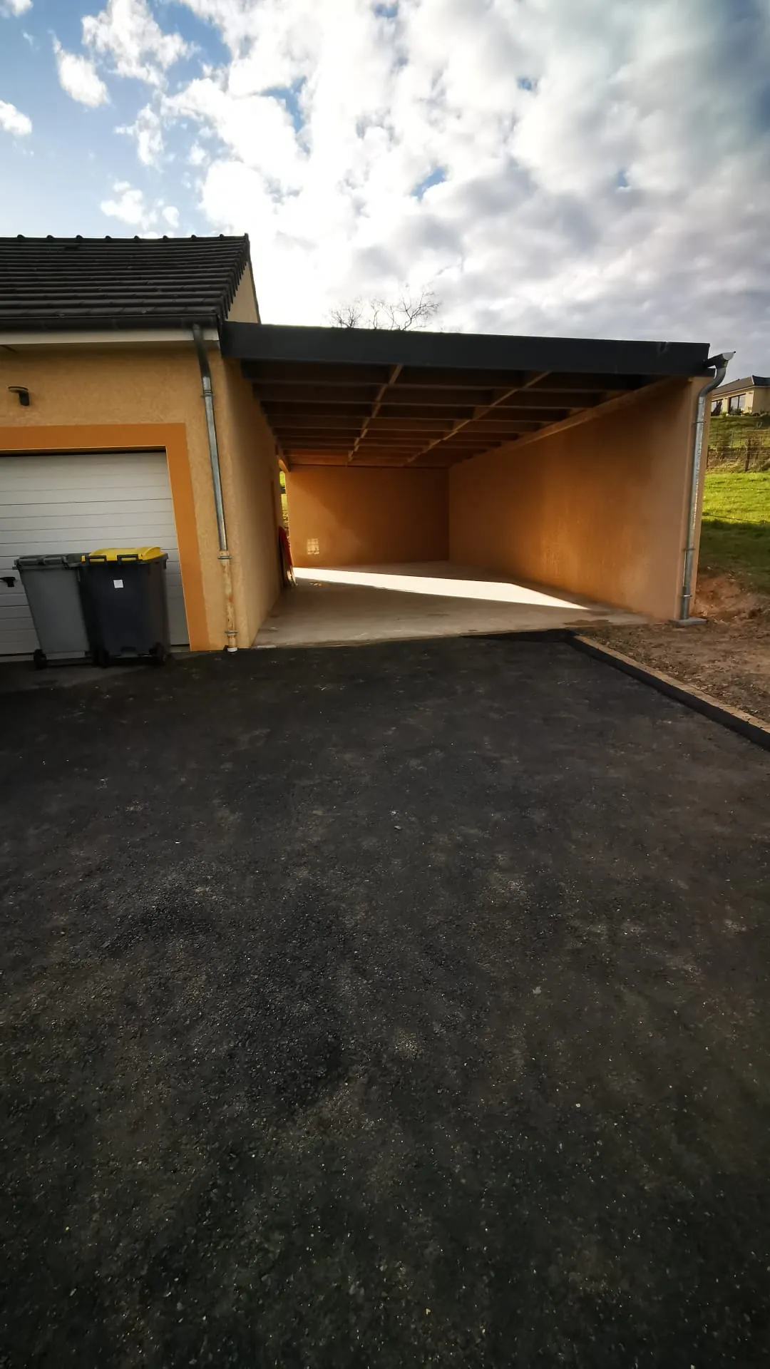 Nouvel enrobé bitumineux noir devant un garage et sous un auvent adossé à la maison.