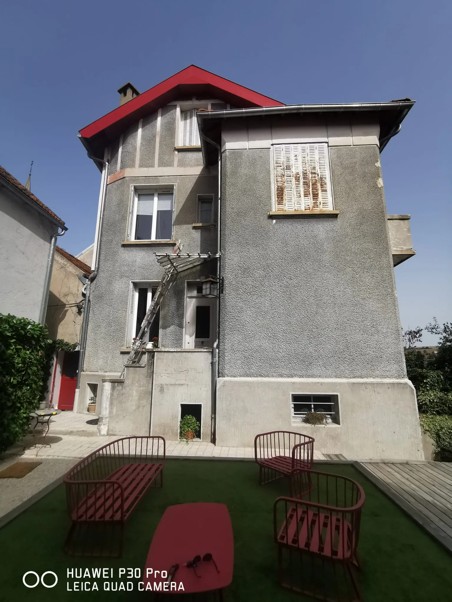 Façade arrière d'une grande bâtisse en crépi gris avec bordures de toit peintes en rouge.
